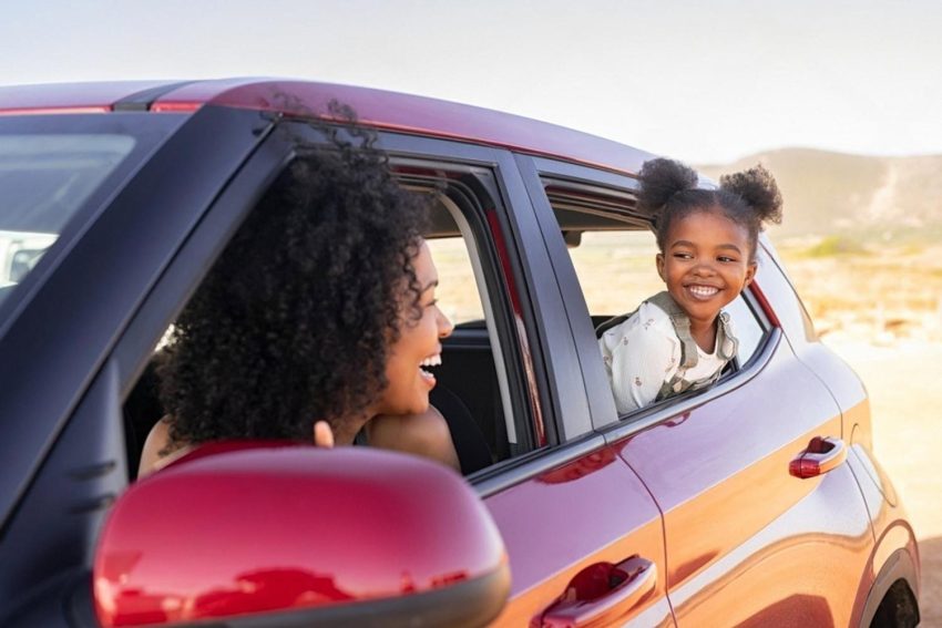 Mother and daughter smiling from the windows of a red family SUV on a road trip